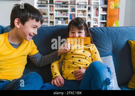 Ragazzo sorridente che tiene l'emoticon sopra il viso della ragazza mentre si siede sul divano a casa Foto Stock