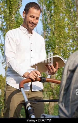 Uomo d'affari sorridente con cuffie intrauricolari che tengono un libro al passeggino del bambino nel parco Foto Stock