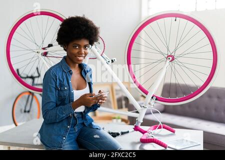 Sorridente giovane donna che usa il telefono cellulare con un ciclo capovolto sul tavolo a casa Foto Stock