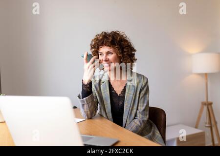 Sorridente freelance che invia messaggi vocali tramite smartphone mentre lavora a casa Foto Stock