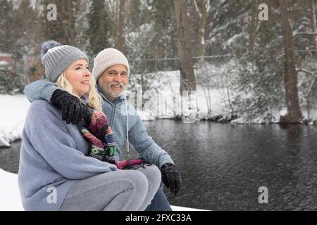 Uomo che abbraccia la donna mentre si siede vicino al lago durante l'inverno Foto Stock