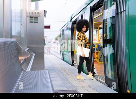 Donna afro che tiene il laptop mentre entra in treno alla stazione ferroviaria Foto Stock