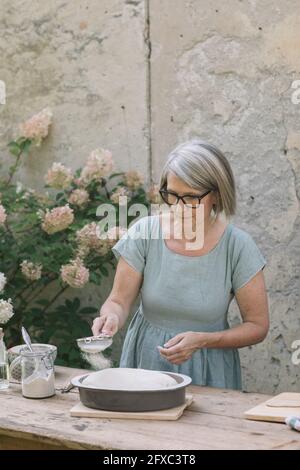 Donna matura che spruzzava zucchero in polvere attraverso il setaccio mentre preparava il pane in cortile Foto Stock