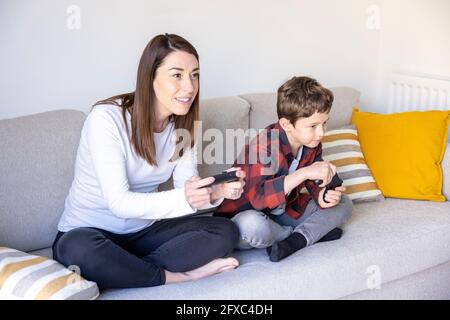 Madre curiosa che gioca a un videogioco di figlio sul divano a casa Foto Stock