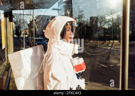 Astronauta femminile in tuta spaziale in piedi alla fermata dell'autobus durante una giornata di sole visto attraverso il vetro Foto Stock