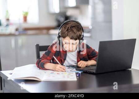 Ragazzo che indossa le cuffie scrivendo sul libro mentre studia a casa Foto Stock