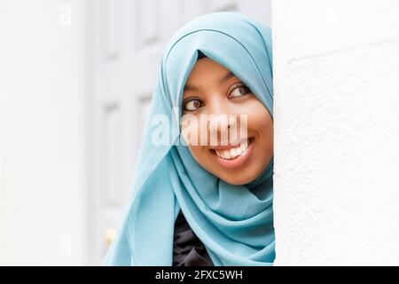 Donna sorridente che indossa hijab sbirciando intorno al muro Foto Stock