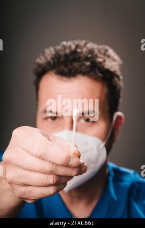 Operatore sanitario di sesso maschile che indossa maschera protettiva viso mostrando il tampone nasale di fronte a sfondo grigio Foto Stock