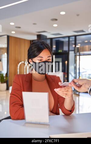 Donna che indossa la maschera facciale che prende la chiave di carta dall'addetto alla reception dell'hotel Foto Stock