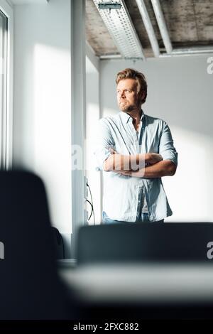 Uomo maturo premuroso con le braccia incrociate che guardano via mentre si è in piedi a casa Foto Stock