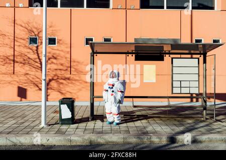 Giovane astronauta femminile in tuta spaziale in attesa alla fermata dell'autobus durante la giornata di sole Foto Stock