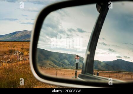 Specchio a vista laterale che riflette il giovane uomo in piedi sulla cima della recinzione guardando attraverso il binocolo alle montagne circostanti Foto Stock