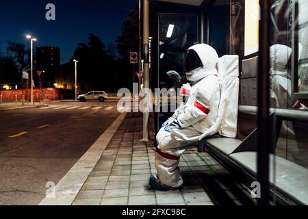 Giovane astronauta femminile in tuta spaziale in attesa alla fermata dell'autobus durante la notte Foto Stock