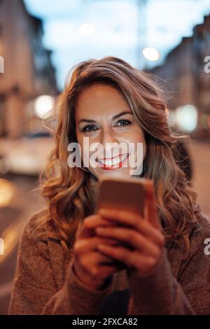 Donna mid adulta sorridente mentre tiene in mano uno smartphone Foto Stock