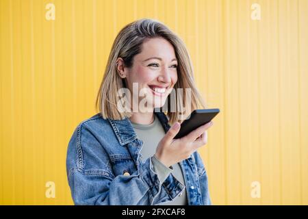 Donna felice che invia la segreteria telefonica tramite smartphone da parete gialla Foto Stock