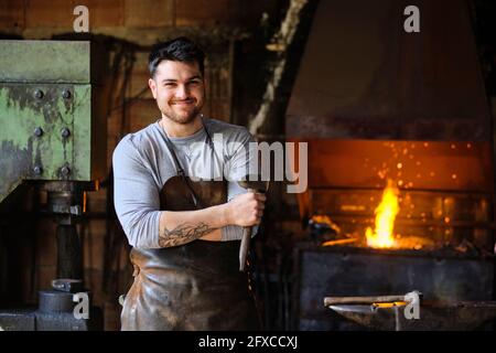 Sorridente giovane fabbro maschio in grembiule in piedi con martello in officina Foto Stock