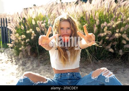 Donna che mangia mela mostrando segno di pace mentre si siede sulla sabbia durante il giorno di sole Foto Stock