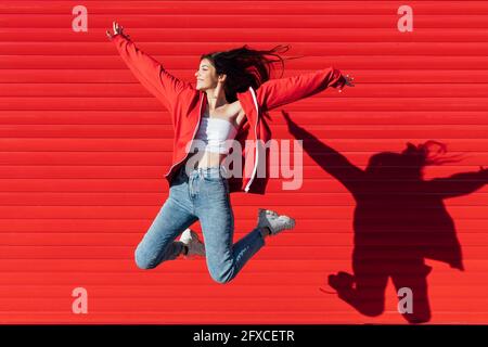 Ragazza adolescente sorridente con le braccia allungate che saltano di fronte al muro rosso Foto Stock