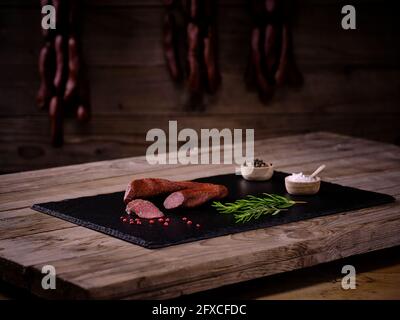 Salsiccia di Landjager con rosmarino, sale e pepe in ciotole e grani di pepe rosso su lastra di pietra ardesia nera Foto Stock