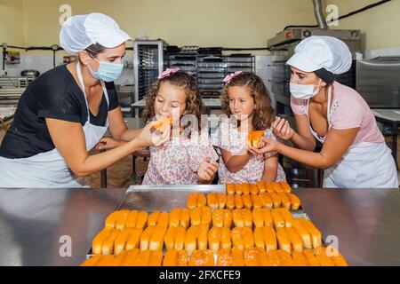 Donne che indossano maschera protettiva che alimenta pasticceria alle ragazze gemelle in cucina panetteria Foto Stock