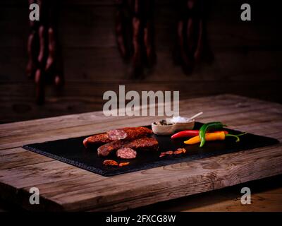 Salsiccia di Landjager con sale e pepe in ciotole, peperoncino e peperoncino fresco su lastra di pietra ardesia nera Foto Stock
