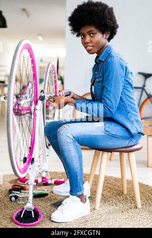 Donna afro acconciatura seduta su sgabello riparando la bicicletta a casa Foto Stock