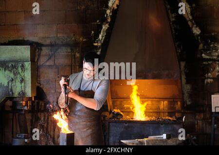 Giovane fabbro maschio che brucia metallo nel fuoco in officina Foto Stock