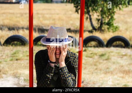 Defocalizzare la giovane donna sorridente in camicia a scacchi e cappello che oscillano sull'altalena sul parco giochi. Donna stanca che ricopre il viso con le mani. Zona di campagna Foto Stock