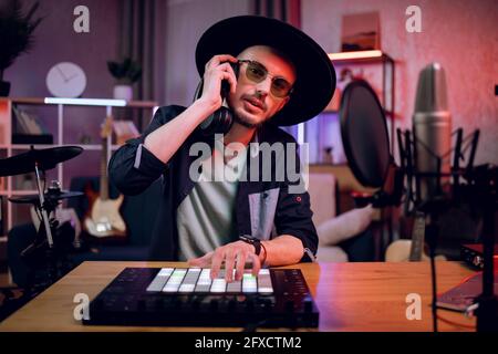 Uomo bearded in cappello e occhiali seduti in camera oscura e mescolando suoni usando il controller dj e le cuffie. Produzione musicale. Processo creativo. Foto Stock