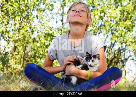 Defocus bambina bionda carezza gatto, nero e bianco gattino piccolo. Natura verde estate sfondo. Ragazza che tiene e stroking animale domestico. Bambini Foto Stock