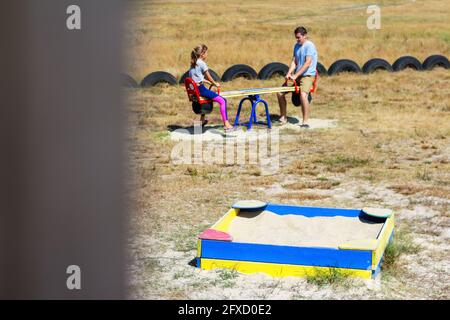 Defocus ragazza oscillare sul campo da gioco con il giovane uomo, ragazzo, fratello maggiore. Campagna. Swing e sandbox blu e rosso brillante. Estate in famiglia Foto Stock