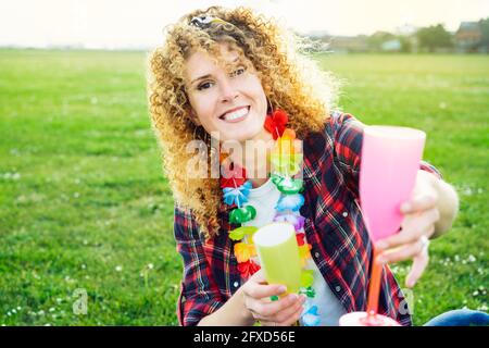 Donna sorridente felice capelli ricci in hawaiana collana dandogli un bicchiere da cocktail per un toast. Festeggia con allegria la vita in un picnic all'aperto in un parco cittadino Foto Stock