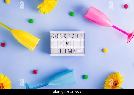 Vista dall'alto piatto di scatola luminosa con iscrizione al tempo del cocktail, bicchieri da cocktail in plastica brillante, gocce di caramelle multicolore e fiori di gerbera gialli su Foto Stock