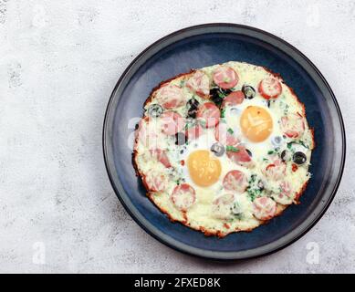 Uova fritte con cipolle verdi, formaggio, salsicce e olive su un piatto rotondo su fondo grigio chiaro. Vista dall'alto, disposizione piatta Foto Stock