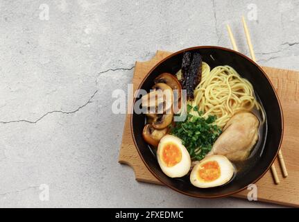 Ramen con pollo, uovo sottaceto, funghi e alghe in una ciotola scura su uno sfondo grigio chiaro. Vista dall'alto, disposizione piatta Foto Stock