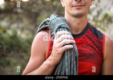 Un arrampicatore maschio di aspetto caucasico con una corda avvolta in mano. Avvolgimento della fune, nodo, stoccaggio delle attrezzature. Mano in magnesia bianca. Rosso sport Jerse Foto Stock