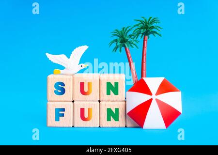 SUN DIVERTENTE scritta fatta di blocchi alfabetici in legno, figurine gabbiano, palme giocattolo e ombrello su sfondo blu. Concetto di vacanza estiva. Foto Stock