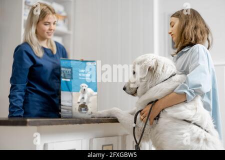 Donna proprietaria con grande cane bianco alla reception in clinica veterinaria scegliendo cibo secco per l'animale domestico. Il cane salì le zampe sul tavolo. Cura e trattamento degli animali domestici Foto Stock