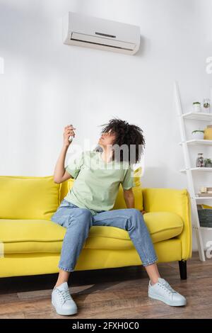 donna afro-americana delusa che tiene il telecomando mentre si siede il divano giallo e sofferente di calore Foto Stock