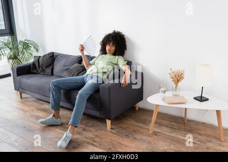 donna afroamericana che agita con la ventola mentre si siede divano grigio e soffriva di calore Foto Stock