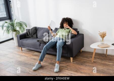 donna afroamericana che agita con la ventola mentre beve acqua e seduto sul divano grigio Foto Stock