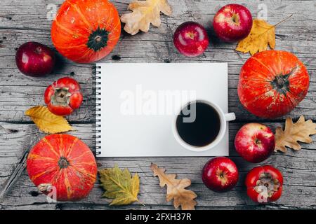 Colazione autunnale Mockup mattina. Taccuino bianco di carta, zucche, mele, persimmons, tazza di caffè e foglie d'autunno secche su sfondo di legno. Foto Stock