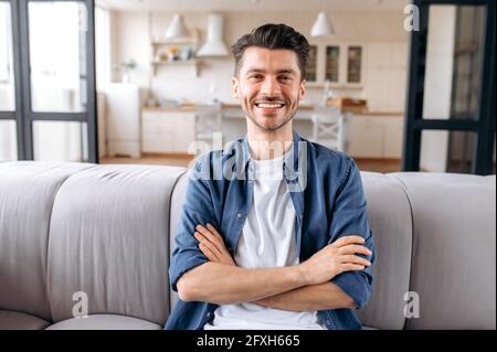 Ritratto di un attraente giovane caucasico, tranquillo e gioioso, seduto a casa con le braccia incrociate sul divano del soggiorno, indossando abiti casual di stile, guardando la macchina fotografica e sorridendo amichevole Foto Stock