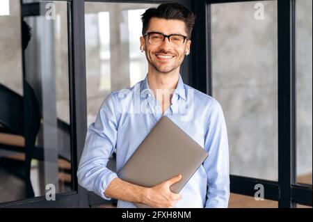 Ritratto di un giovane e attraente uomo d'affari caucasico o lavoratore d'ufficio, indossando camicia e occhiali, in piedi in un moderno ufficio con computer portatile, guardando la fotocamera e sorridendo amichevole Foto Stock