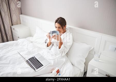 Affascinante giovane donna che passa il tempo a letto al computer portatile e gustoso pasto in casa appartamento Foto Stock