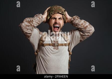 Furioso uomo militare in casco urlando e afferrando la testa isolato su sfondo nero Foto Stock