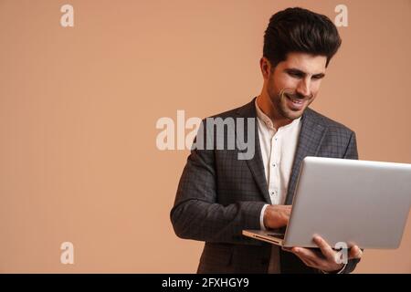 Sorridente giovane in giacca in piedi mentre si lavora con il computer portatile isolato su sfondo beige Foto Stock