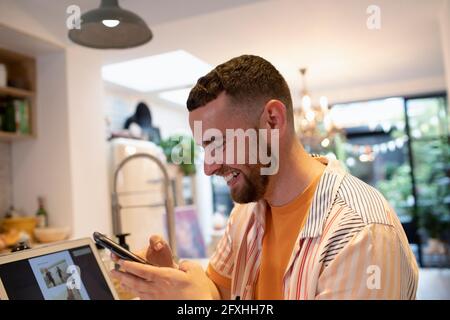 Felice giovane uomo che usa lo smartphone al computer portatile in cucina Foto Stock