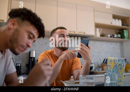 Felice coppia gay maschio utilizzando smartphone e mangiare in cucina Foto Stock