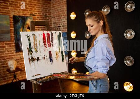 Pittore dello studio che crea un quadro astratto su tela utilizzando dipinti ad olio e coltello a palette. Giovane artista femminile che lavora. Processo creativo, relax Foto Stock